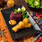 A vibrant Indonesian-inspired dish is artfully plated on a black rectangular plate. Two skewers of golden grilled tofu satay rest beside a mound of deep purple riceberry coconut rice, garnished with fresh cilantro and lime wedges. A dollop of creamy peanut sauce with a hint of chili sits nearby. The plate is set atop colorful Indonesian batik fabrics, with a bowl of fresh green lettuce and bright red chilies in the background, creating an inviting and exotic presentation.
