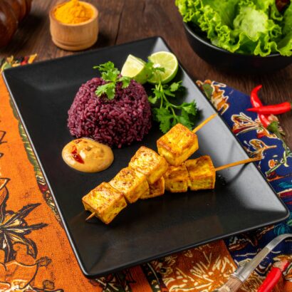A vibrant Indonesian-inspired dish is artfully plated on a black rectangular plate. Two skewers of golden grilled tofu satay rest beside a mound of deep purple riceberry coconut rice, garnished with fresh cilantro and lime wedges. A dollop of creamy peanut sauce with a hint of chili sits nearby. The plate is set atop colorful Indonesian batik fabrics, with a bowl of fresh green lettuce and bright red chilies in the background, creating an inviting and exotic presentation.