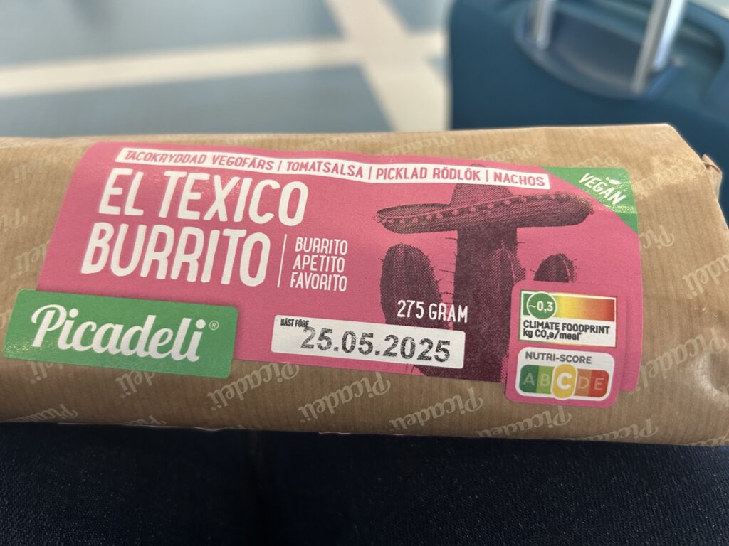 Picadeli El Texico Burrito package showing a brown paper wrapper with pink and green branding. The 275-gram vegan burrito contains spiced veggie mince, tomato salsa, pickled red onion, and nachos, with a best before date of May 25, 2025. The package prominently displays a climate footprint label showing 0.3 kg CO2e per meal in dark green, indicating a very low carbon footprint. A Nutri-Score rating of C is visible in the yellow section of the five-color nutritional rating scale (A-E), positioned on the lower right of the packaging.
