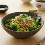 A bowl of soba noodles topped with fresh broccoli florets, edamame beans, sliced green onions, and shredded nori seaweed, served in a dark speckled ceramic bowl. Two pieces of nori sheet are artfully placed upright in the dish as garnish. The bowl sits on a light wooden surface alongside chopsticks, small dipping sauce bowls, and a traditional tea cup in the background, creating an authentic Japanese dining presentation.