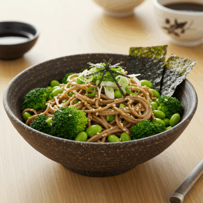 A bowl of soba noodles topped with fresh broccoli florets, edamame beans, sliced green onions, and shredded nori seaweed, served in a dark speckled ceramic bowl. Two pieces of nori sheet are artfully placed upright in the dish as garnish. The bowl sits on a light wooden surface alongside chopsticks, small dipping sauce bowls, and a traditional tea cup in the background, creating an authentic Japanese dining presentation.